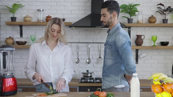 Joyful Handsome Young Man Talking to Beautiful Woman Cooking Salad in Kitchen and Giving Envelope