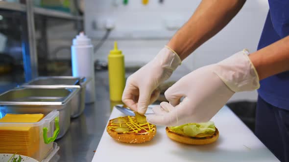 Hands in latex gloves put pickles on top of fried bun. Making burger on white table.