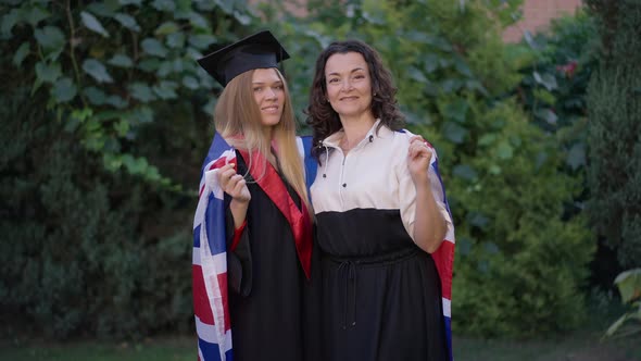 Medium Shot Proud Mature Mother and Young Graduate Daughter Posing with British Flag Outdoors on