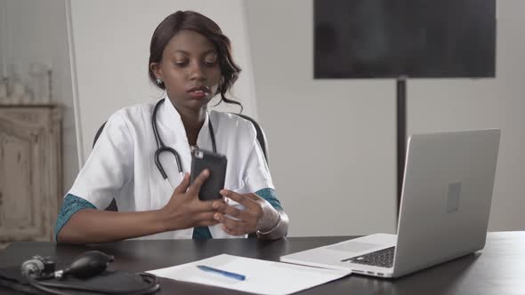 Black Female Doctor Sits at an Office Desk Looking To Camera, African American Nurse Work on Laptop