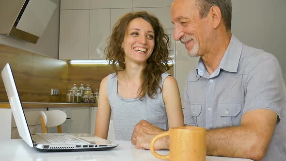 Attractive Young Daughter and Senior Father are Using Laptop Sitting in Modern Kitchen in Apartment