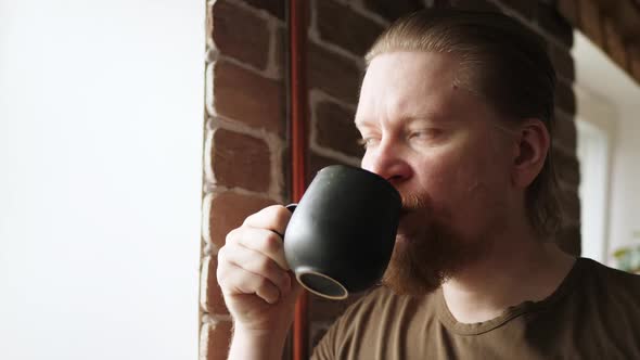 Stern Redbearded Man Drinking From Black Mug By Window Against Brick Wall