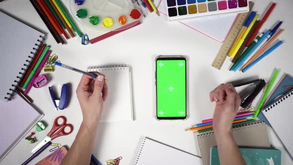 Man Swipes Up and Zooms on a Smartphone with Green Screen Lying on the White Desk Among School Tools alt