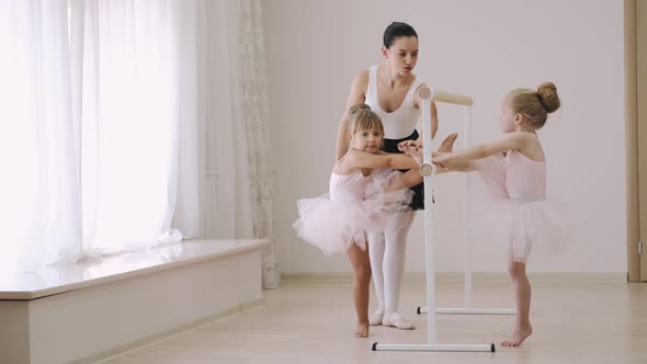 Two Little Girls Practicing Choreographic Elements on Ballet Barre with Help of Teacher in Dance