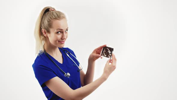 Young Blond Cheerful Female Gynecologist in Blue Uniform Showing Pregnancy USG Picture to the Camera