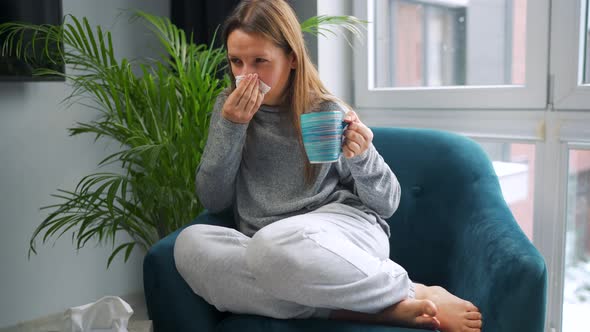 Unhealthy Woman Sits in a Chair Drinks Hot Tea or a Cold Medicine and Sneezes or Blows Her Nose Into
