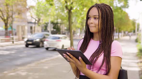 A Young Black Woman Works on a Tablet with a Smile in the Street in an Urban Area