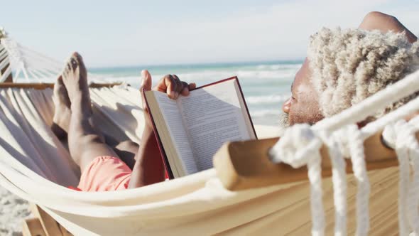 Senior african american man reading and lying in hammock on sunny beach