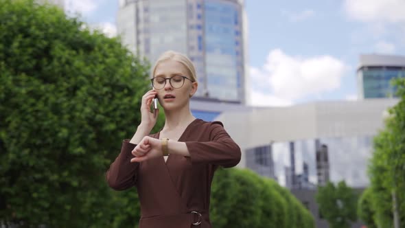 Serious Busy Woman Looks at Her Watch and Makes an Appointment By Phone Against the Backdrop of a