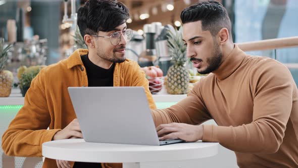 Two Arab Men Sit Looking at Laptop Screen Communicate Smiles Studying Using New Computer Application