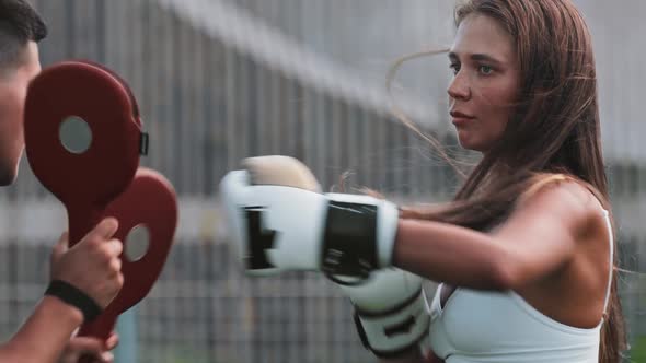 Boxing Training  Young Woman with Long Hair Punching the Mitts on the Hands of Her Male Trainer