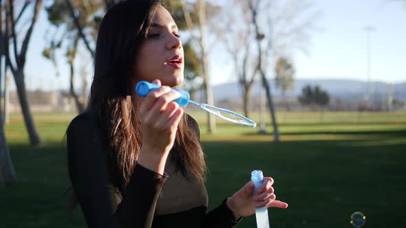 A happy young woman blowing iridescent bubbles and being playful and smiling outdoors in sunshine.