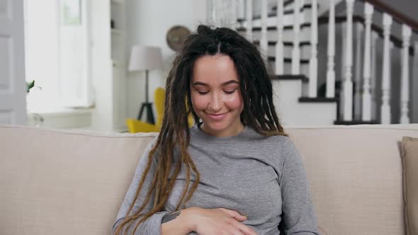 Pregnant Girl with Dreadlocks Sitting on the Sofa at Home, Caressing Her Tummy