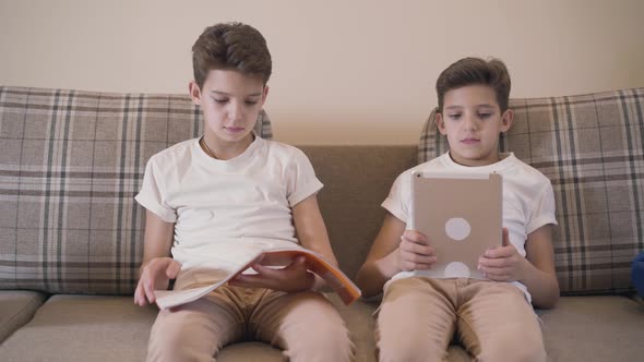 Portrait of Young Caucasian Schoolboy Sitting with Notebook at the Couch As His Twin Brother Showing