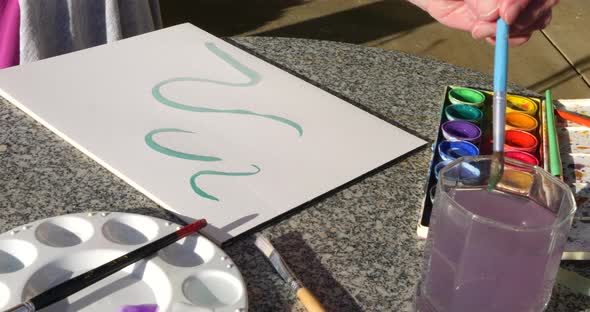 A woman artist painting an abstract green swirl with watercolor on a table in an outdoor art studio.