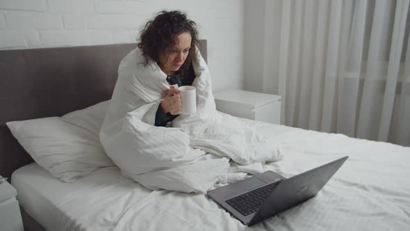 Sick Woman Drinking Hot Beverage Watching Video on Laptop Indoors