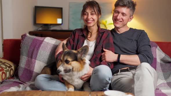 Happy Couple with Cute Pet Welsh Corgi Dog Sitting on Couch at Home at Evening