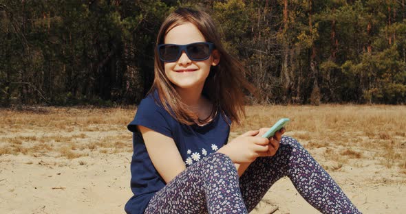 Cute Little Girl Holds Phone in Hand and Sits in Summer Forest