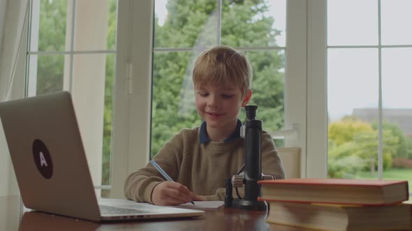 Young Boy Looking at Microscope and Talking to Laptop Studying Online From Home