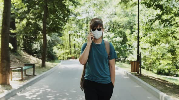 Young Man with a Guitar on the Back Is Walking in the Park and Talking on Phone