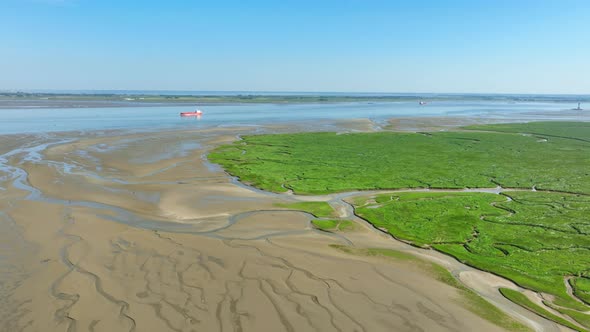 Aerial shot of wetlands leading into a river where a cargo ship is navigating its way under a blue s