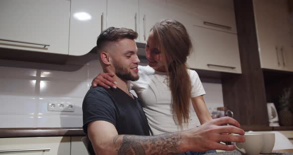 Girl Sits On The Guy's Hands At The Table At Home In The Kitchen