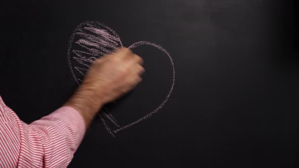 A man's hand in a red and white striped shirt draws a heart with pink chalk on a black board