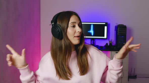 Young Woman in Pink Sweatshirt in Headphones Dancing and Singing in Modern Room with Neon Background