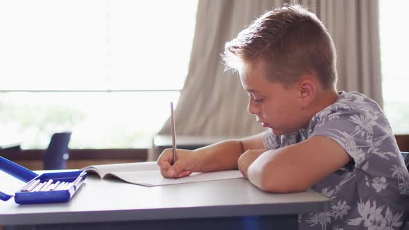 Portrait of happy caucasian schoolboy sitting at classroom, making notes, looking at camera