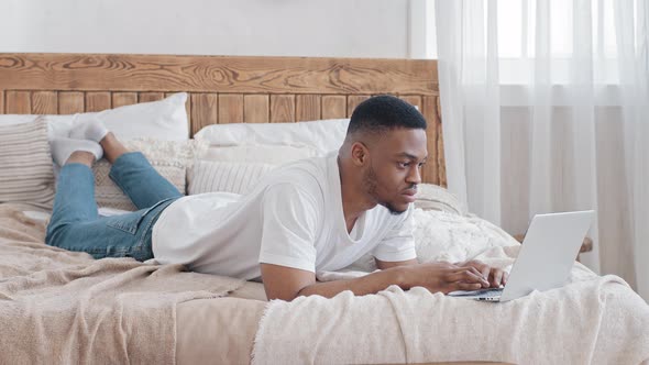 Young Handsome African Man Freelancer Working Online Lying on Bed Uses Laptop