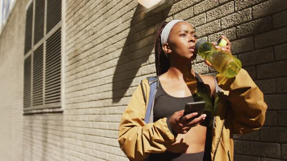 African american woman exercising outdoors drinking water and using smartphone in the city