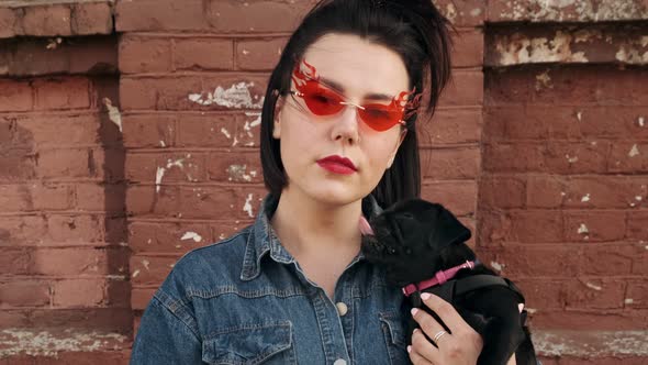 Stylish woman in fashionable red glasses looks at the camera with a black dog pug puppy