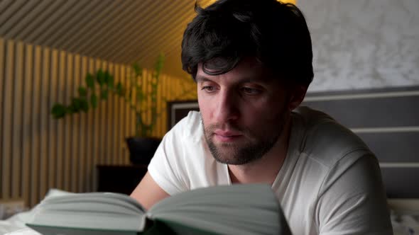 Man Reads a Book at Home in the Evening on the Bed