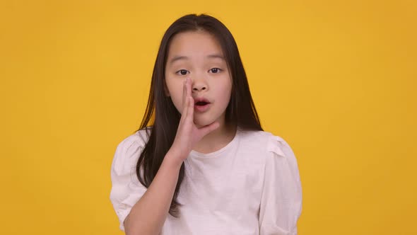 Cute Asian Girl Whispering Secret To Camera Covering Her Mouth with Hand Orange Studio Background