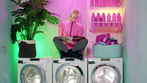 Nice Punk Woman with Short White Hair and Pink Jumpsuit Sits on Washing Machines in Laundry Room and
