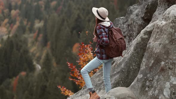Happy Woman Hiker in Hat Stands with Rised Hands on Edge of Cliff and Looks Around Enjoying Nature