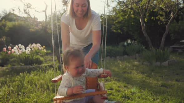 A smiling mother swings a swing with her petulant child.