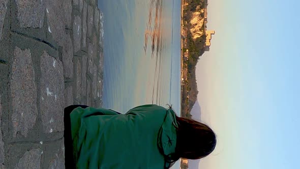 Back view of woman sitting on jetty facing lake and castle. First-person view and VERTICAL format