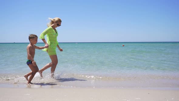 Young Woman Runs Along the Shore of the Beautiful Azure Sea, Holding a Child By the Hand.