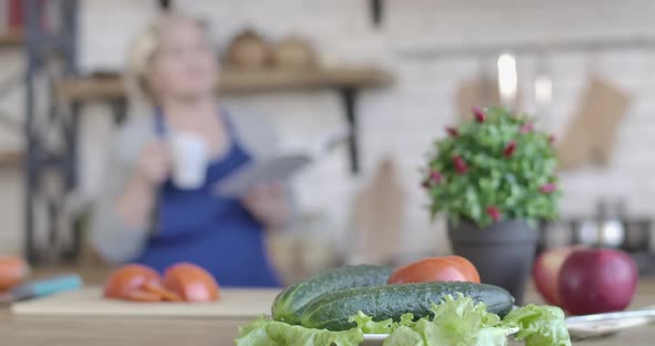 Close-up of Fresh Vegetables Lying on Table in Kitchen As Blurred Woman Drinking Coffee and Reading