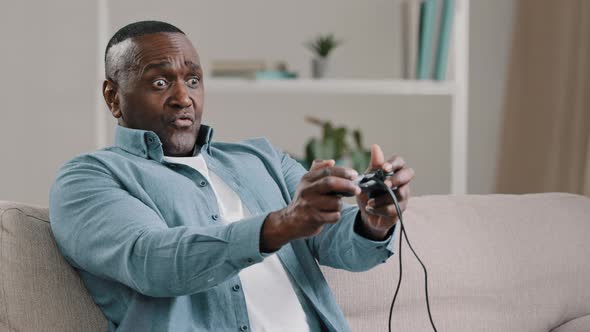Mature African American Man Sitting on Couch in Room Attentively Playing Video Games on Console