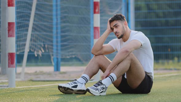 Frustrated Upset Brooding Middle Eastern Footballer Sitting on Grass of Soccer Field Against Goal