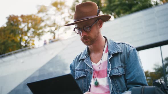 Portrait of Young Bearded Hipster Man and in Hat Sits on Bench in City Center in Park Using Laptop