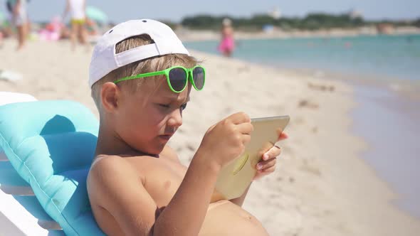 Boy Sitting By the Sea Uses a Tablet on a Sunny Summer Day