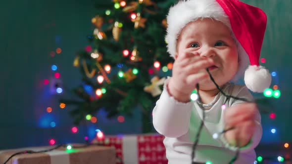 Portrait of a funny baby girl in a Christmas hat, playing with festive lights