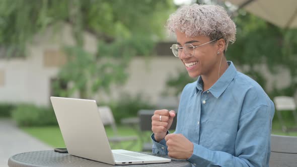 Young African Woman Celebrating Success on Laptop in Outdoor Cafe