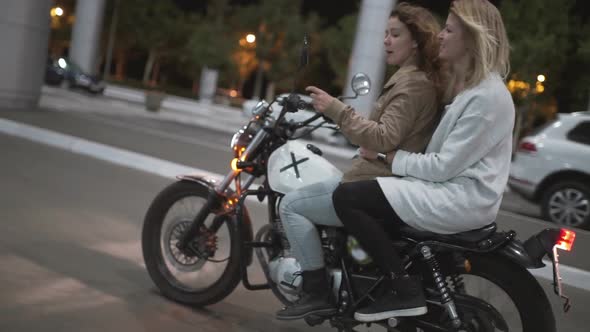 Two Beautiful Young Women Riding a Motorcycle at Night on City Street and Talking to Each Other