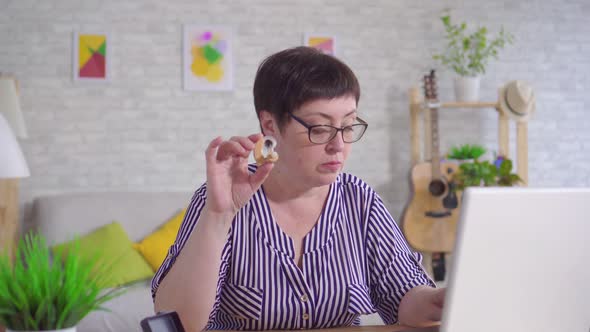 Woman with Glasses Sitting at a Table in the Living Room Holding a Hearing Aid Uses a Laptop