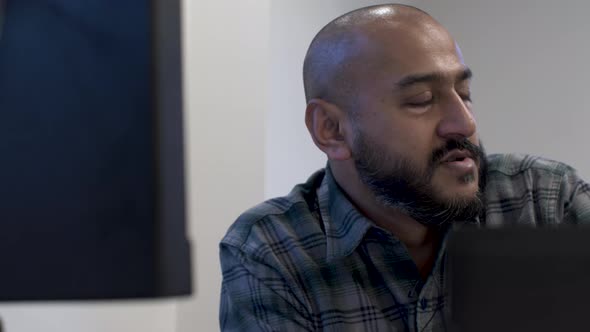 Ethnic Minority Bald Male Wearing Green Cheque Shirt Talking To Colleague Across From Him. View Behi