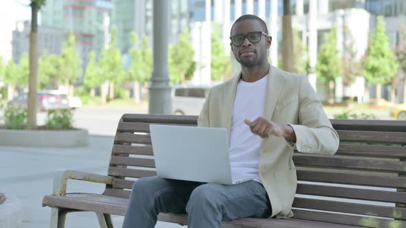 African Man with Laptop Showing Thumbs Down While Sitting Outdoor on Bench
